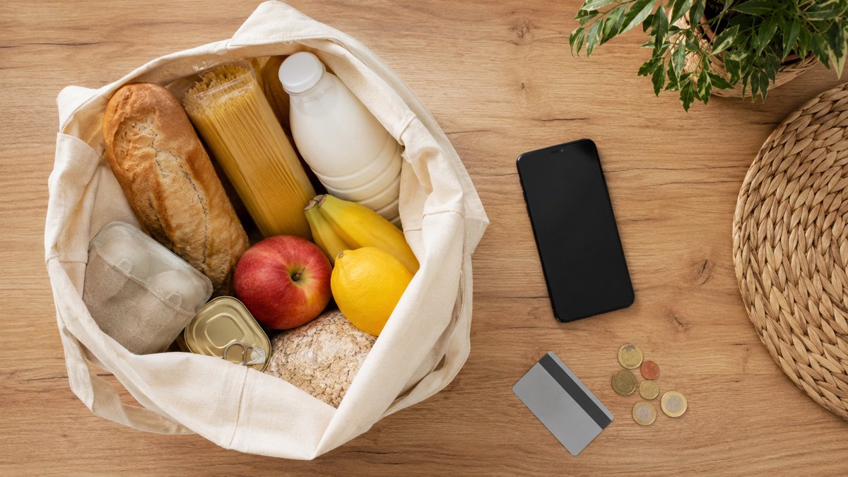 Bolsa con alimentos básicos (pan, leche, fruta, pasta) junto a móvil, tarjeta y monedas en una mesa